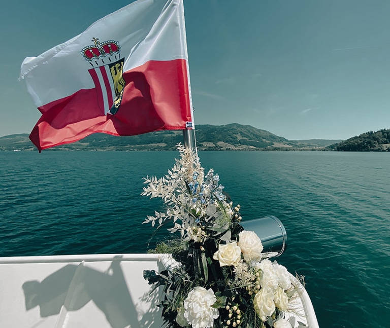 Die Flagge mit dem oberösterreichischen Wappen weht am Bug des Passagierschiffes MS Mondseeland. Ein Blumengesteck mit weißen Rosen und grünem Blattwerk schmückt den Schiffsbug. Man blickt über die Fahne hinweg auf die Wasseroberfläche des Mondsees und die Landschaft.