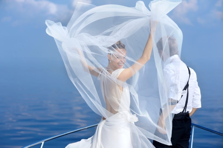 Ein Brautpaar am Bug eines Schiffes. Die Braut lässt den Schleier fliegen und blickt lächelnd zurück.