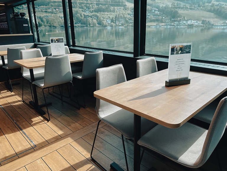 In der Panoramakabine des Passagierschiffes MS Mondseeland stehen zwei Tische mit jeweils grau gepolsterten Stühlen in der Sonne. Auf den Tischen stehen Speisekarten. Durch die Fenster sieht man auf die Wasseroberfläche.