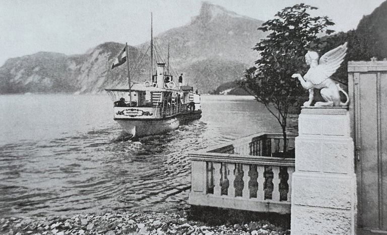 Ein Schwarz-Weiß-Foto des Passagierdampfschiffs Habsburg auf dem Mondsee von hinten circa im Jahr 1890. Im Vordergrund eine Steg mit Balustrade mit einer geflügelten Sphinx auf einer Säule. Im Hintergrund der Schafberg