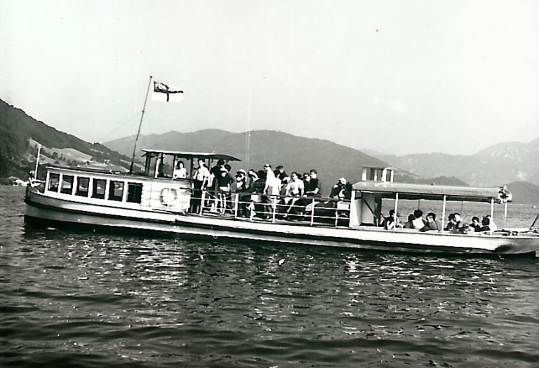 Die MS Helene fährt auf dem Mondsee Ein Schwarzweiß-Foto, das um 1960 aufgenommen wurde. Mehrere Passagiere sind an Deck.