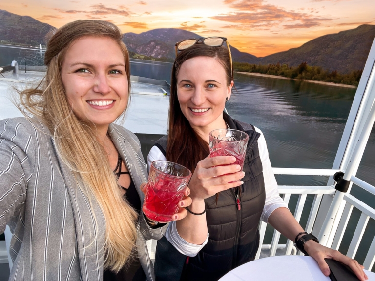 Zwei junge Frauen trinken einen roten Cocktail auf dem Sonnendeck des Passagierschiff MS Mondseeland. Eine ist blond, die andere brünett mit Sonnenbrille im Haar. Beide lachen und machen ein Selfie. Die Sonne geht unter.