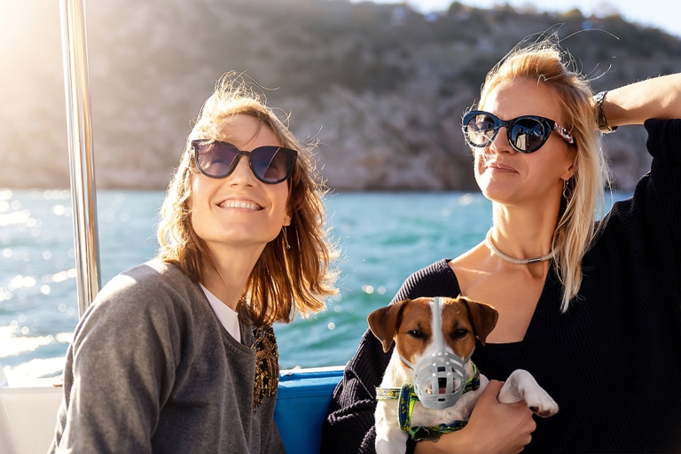 Zwei junge Frauen sitzen mit einem Jack-Russel-Terrier in einem Boot. Es scheint die Sonne. Im Hintergrund sieht man Wasser und Ufer. Der Hund wird von der blonden Frau rechts im Arm gehalten. Beide lachen und tragen dunkle Sonnenbrillen. Der Hund hat einen Maulkorb.