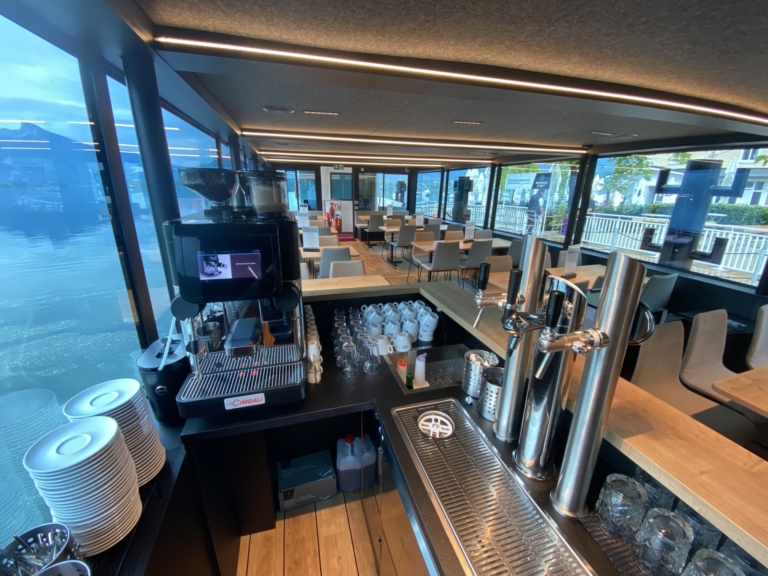 Die moderne mit Holz und LED-Licht ausgestattete Kabine des Passagierschiffs MS Mondseeland. Man blickt ins Innere der kleinen Schiffsbar mit Teller, Kaffeemaschine, Tassen, Gläsern und Zapfhähnen. Die Decke hat Leuchtstreifen.
