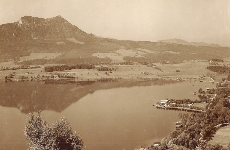 Blick vom Mondseeberg über den Mondsee und die Seevilla auf den Schober. Ein Schwarzweiß-Foto aus dem frühen zwanzigsten Jahrhundert. Es ist Sommer und der See liegt ganz ruhig.