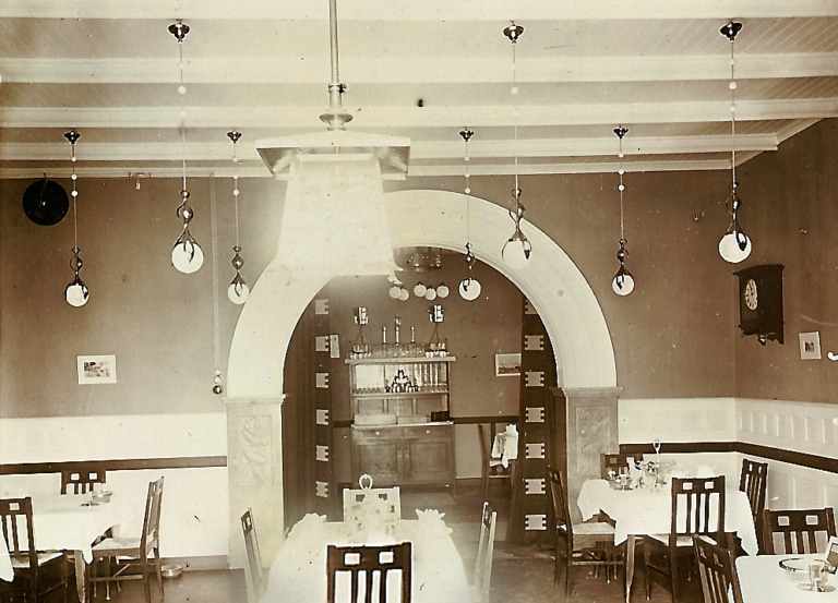 Ein Schwarzweiß-Foto zeigt den Gastraum der Seevilla in Mondsee in den Zwanziger Jahren. Der Stil ist Jugendstil und Art Deco. Es hängen viele kleine Kugellampen von der Decke. Im Mittelpunkt ist ein großer Durchgangsbogen in einen anderen Raum.