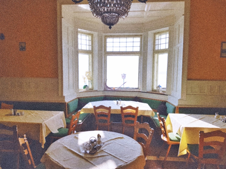 Ein verblichenes Farbfoto zeigt den Gastraum der Seevilla in Mondsee in im Stil der Sechziger Jahre. Der Stil ist eine Mischung aus originalem Jugendstil und jüngerer rustikalen Bestuhlung. Die Wände sind gelb gestrichen. Im Mittelpunkt ein Erker mit drei Fenstern. Die Tische haben weiße Tischdecken.