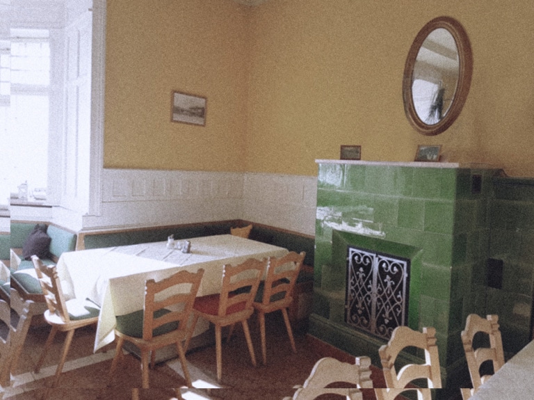 Ein verblichenes Farbfoto zeigt den Gastraum der Seevilla in Mondsee in im Stil der Sechziger Jahre. Der Stil ist eine Mischung aus originalem Jugendstil und jüngerer rustikalen Bestuhlung. Die Wände sind gelb gestrichen. Im Mittelpunkt ist ein schlichter grüner Kachelofen mit Metallgitter und darüber ein ovaler Spiegel.