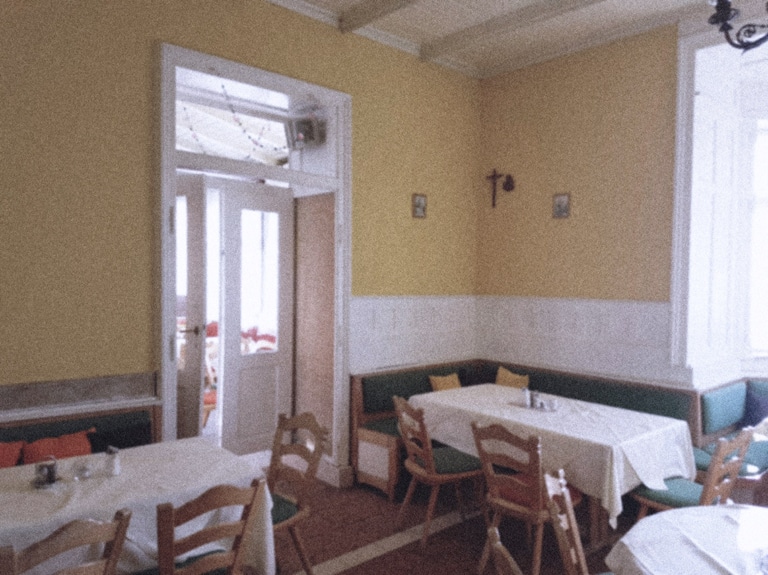 Ein verblichenes Farbfoto zeigt den Gastraum der Seevilla in Mondsee in im Stil der Sechziger Jahre. Der Stil ist eine Mischung aus originalem Jugendstil und jüngerer rustikalen Bestuhlung. Die Wände sind gelb gestrichen. Im Mittelpunkt eine weiße doppelflügelige Türe mti Glaseinsatz. Die Tische haben weiße Tischdecken.