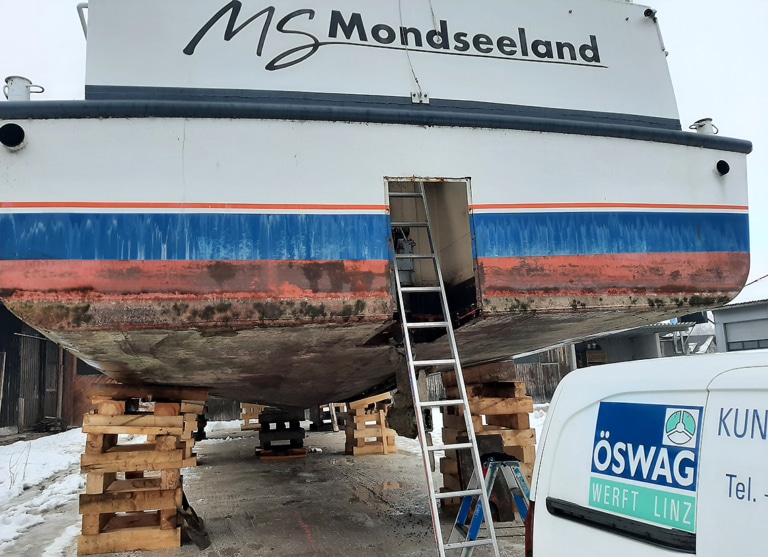 Das Passagierschif MS Mondseeland wird im Winter 2021 auf 2022 renoviert. Es liegt aufgebockt auf Holzbalken im Trockendock. Eine Leiter führt im Heck durch eine Öffnung auf das Deck. Die Lackierung sieht sehr alt aus.