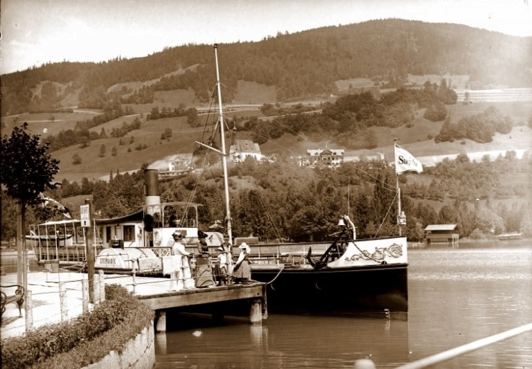 Das Dampfschiff Stephanie liegt am Steg der Seepromenade in Mondsee. Ein Schwarzweiß-Foto, das um 1900 aufgenommen wurde. Mehrere Frauen in historischen Kleidern warten am Steg.