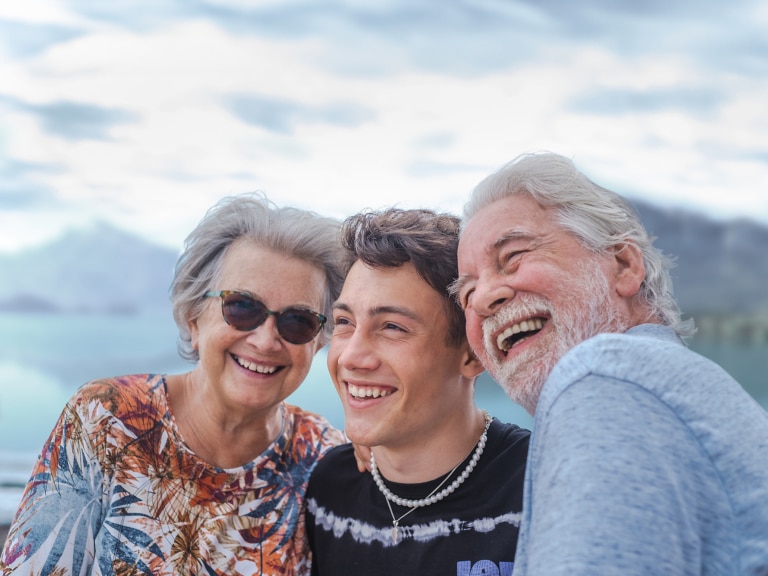 Oma und Opa mit ihrem Teenager Enkelsohn an einem See. Der Junge ist in der Mitte und umarmt seine Großelter. Alle lachen und freuen sich.