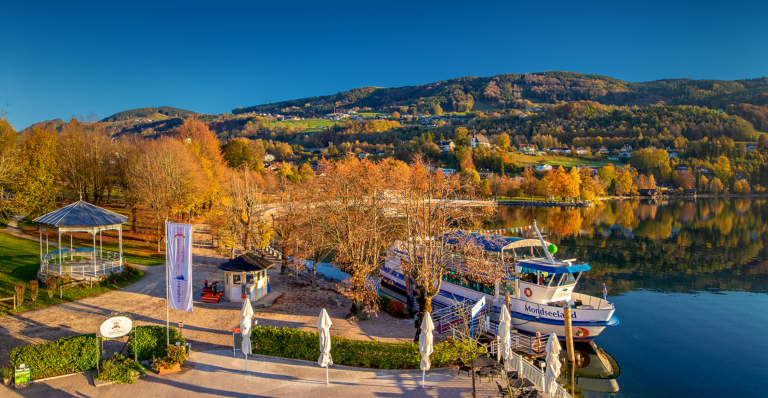 Blick auf die Seepromenade im Herbst. Das Laub der Bäume ist in feurigem Gelb verfärbt. Links sieht man den Musikpavillon und rechts liegt das Passagierschiff MS Mondseeland am Steg.