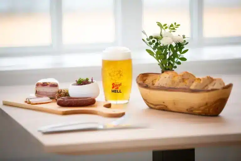Ein gedeckter Tisch des Wirtshauses Mondfleckerl an der Seepromenade Mondsee. Man sieht ein Holzbrett mit einer Jause, Speck und Wurst, sowie einen Brotkorb und ein Bier.